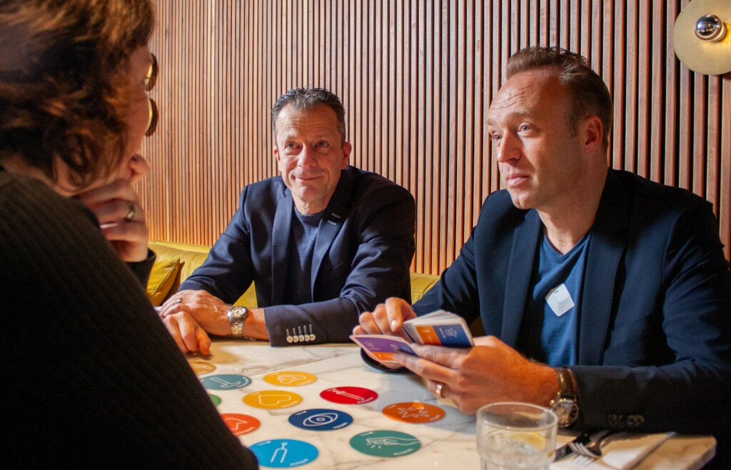 Drie professionals in gesprek aan een ronde tafel met een kleurrijk geheel aan symbolen, gericht op organisatieontwikkeling in samenhang gebruikmakend van het coherentie gedachtengoed.