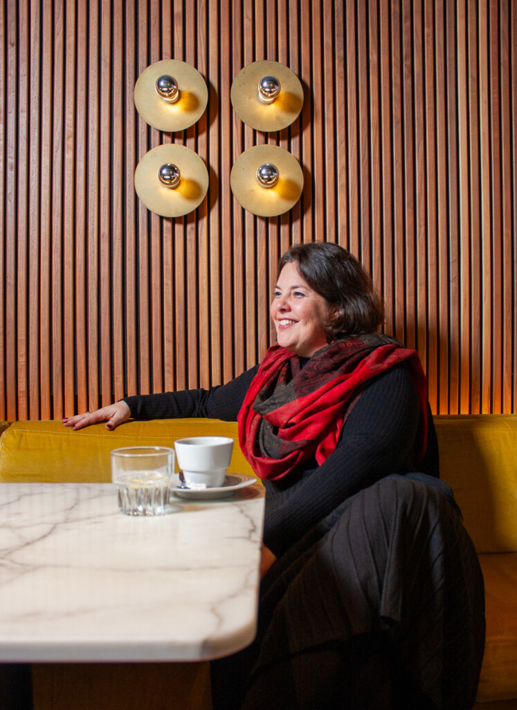 Vrouw zittend aan een marmeren tafel met een kopje koffie, lachend in een café met houten wand en gouden lampen.